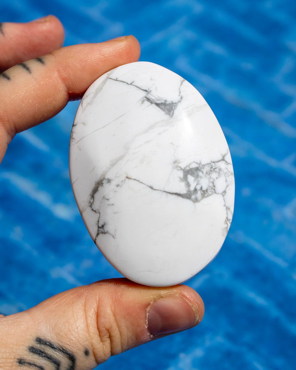 A hand holds a smooth, polished white stone with gray and black marble-like veining against a bright blue textured background.