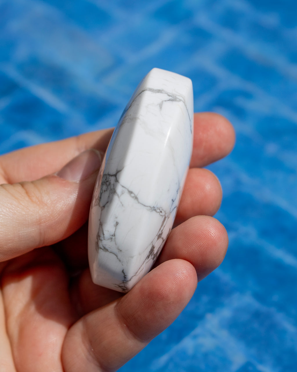 A hand holding a polished white stone or crystal with gray veining, set against a bright blue water background.
