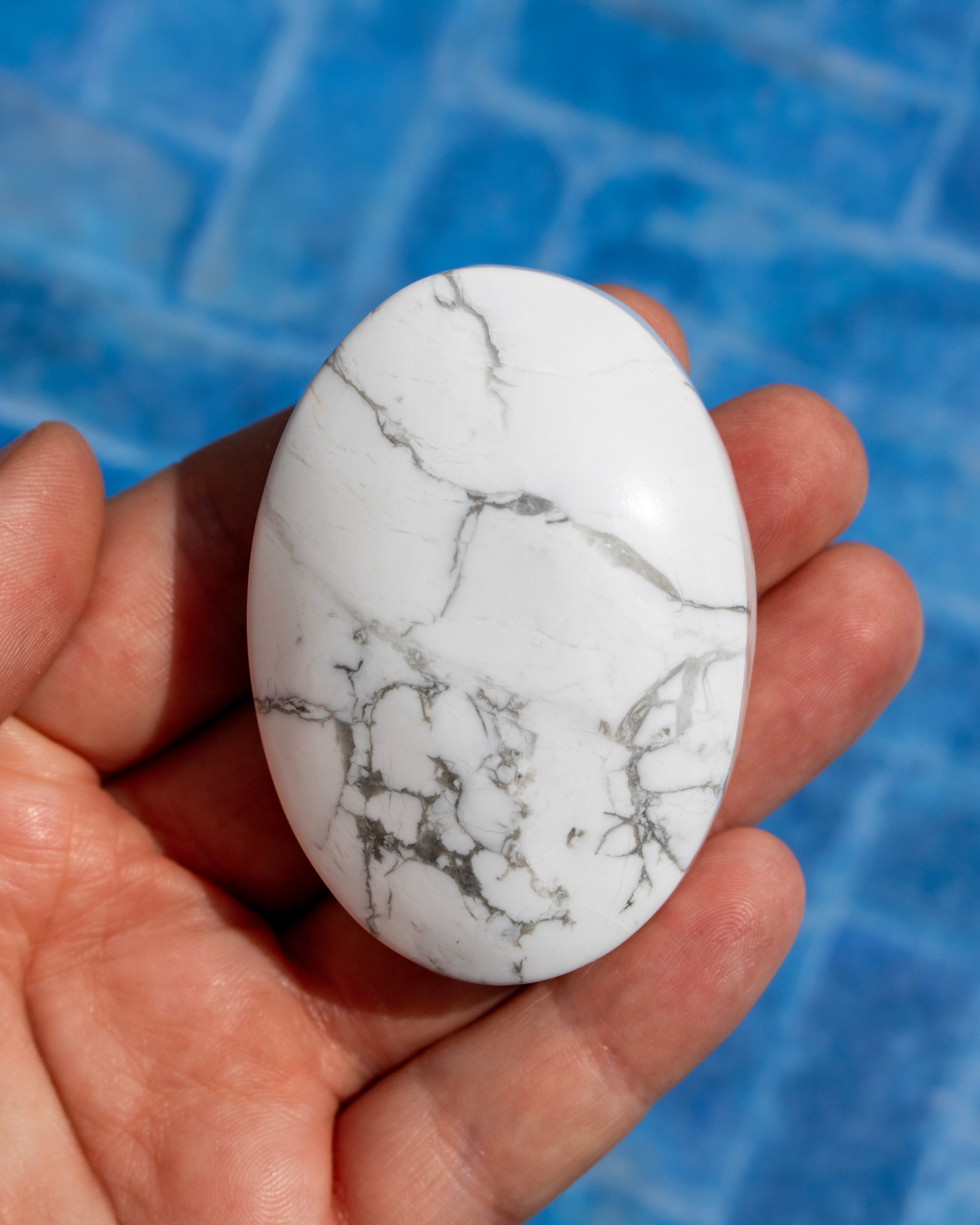 A hand holding a polished white oval stone with gray and black marble-like veining, set against a blue tiled background.