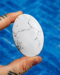 A hand holds a smooth, oval-shaped white stone with natural gray and black veining, set against a bright blue tiled background.
