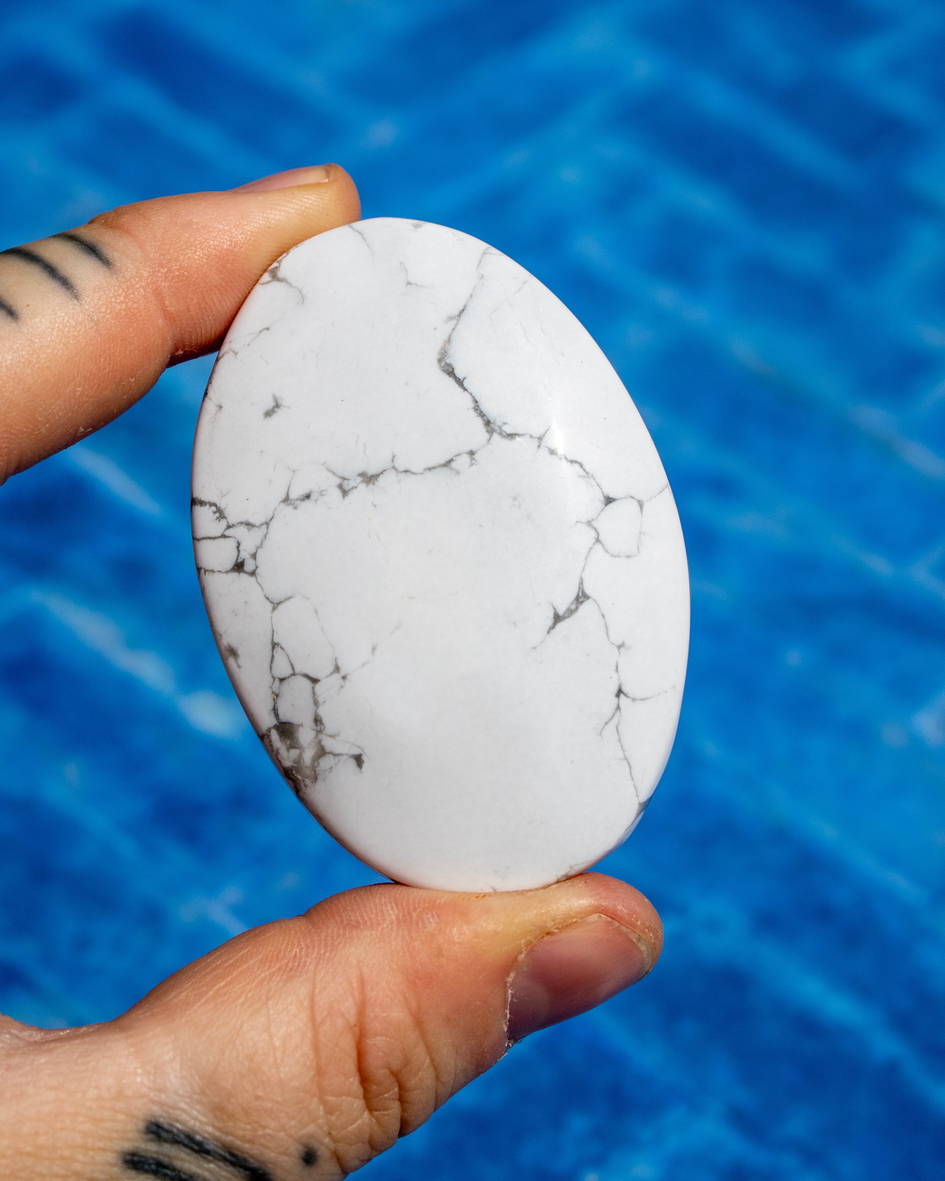 A hand holds a smooth, oval-shaped white stone with natural gray and black veining, set against a bright blue tiled background.