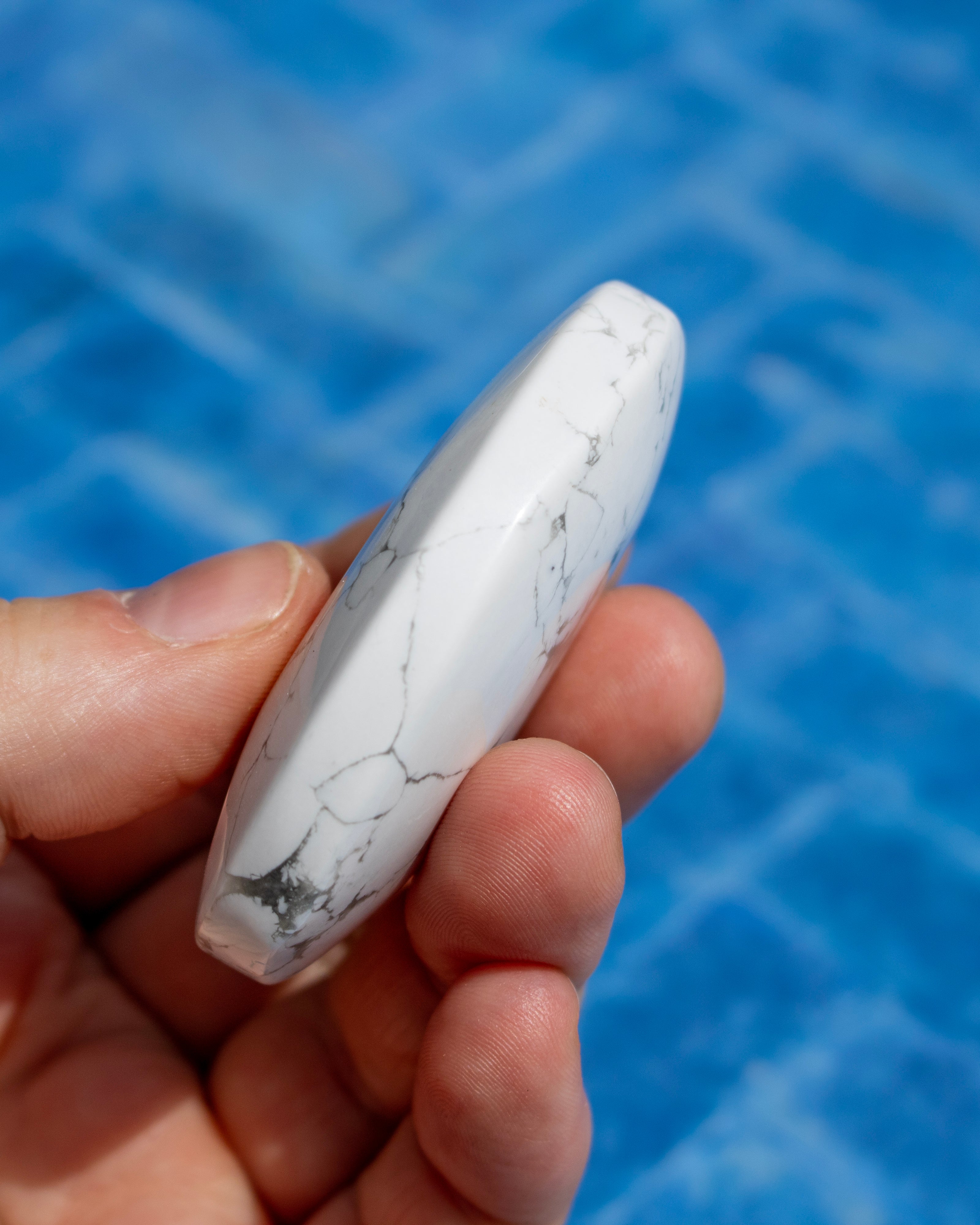 A hand holding a polished white stone cabochon with natural gray veining, set against a blurred blue tiled background.