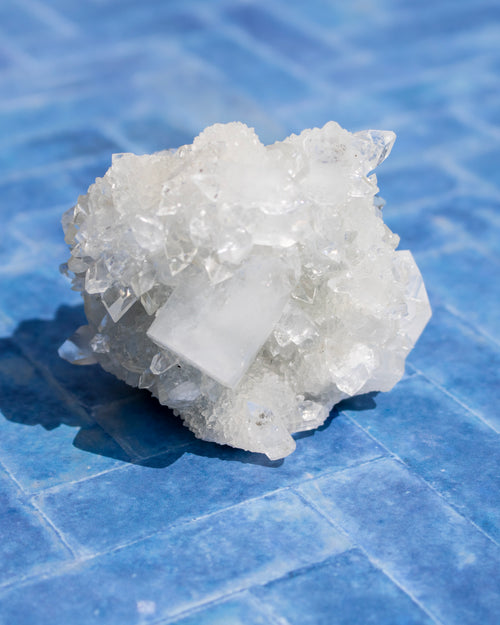 A raw white crystal cluster with a prominent square face, resembling fluorite, resting on a blue tiled surface.