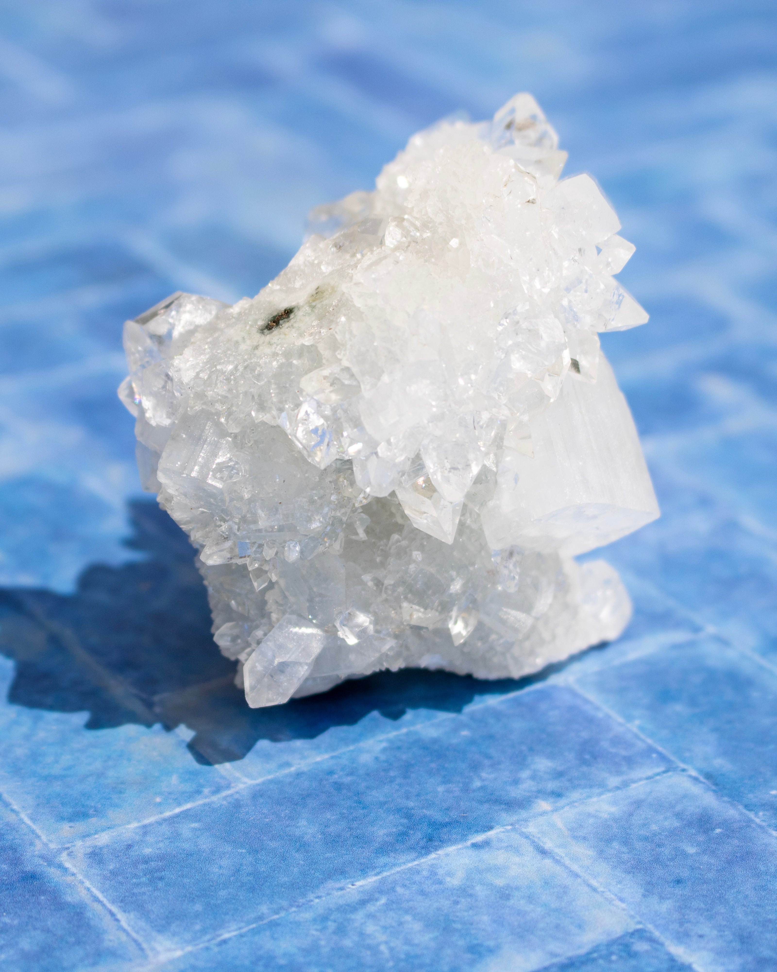 A raw white crystal cluster with sharp faceted mineral points, photographed standing on a blue tiled surface.