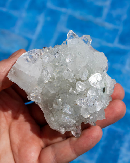 A hand holding a cluster of clear, translucent quartz crystals against a bright blue tiled background.