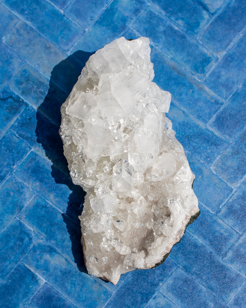 A raw crystal cluster with translucent white and light gray quartz-like facets, photographed from above on a bright blue tiled surface.