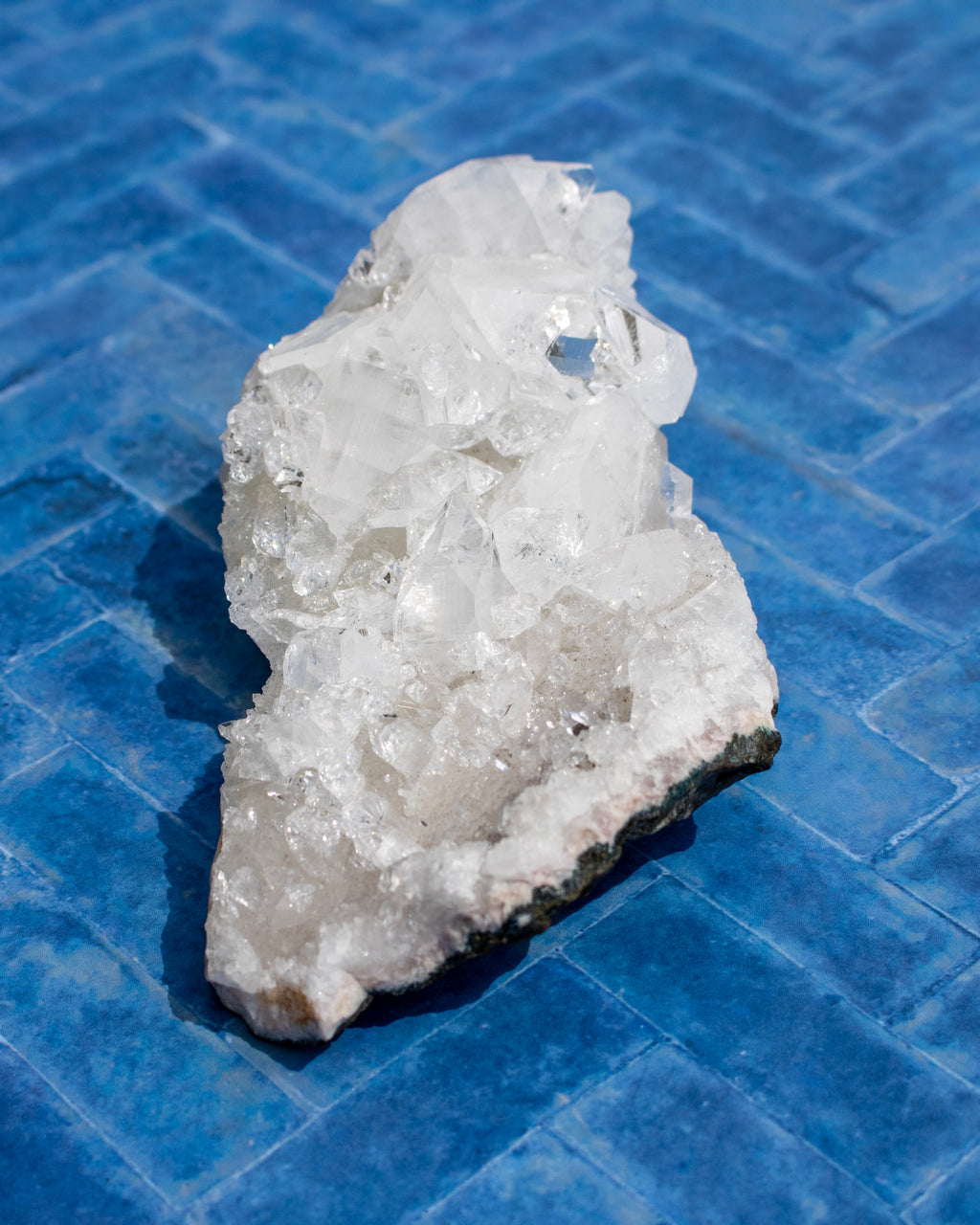A raw crystal cluster specimen with clear, faceted white mineral formations on a rough, dark gray to black matrix base, photographed on a blue tiled surface.
