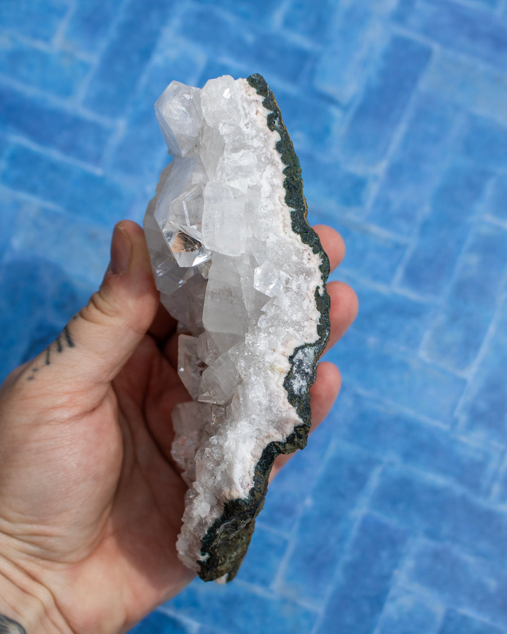 A hand holding a raw crystal cluster featuring large clear quartz crystals on a dark, rough matrix, set against a blue tiled background.