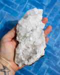 A hand holding a large, rough white crystal cluster with clear faceted mineral pieces, set against a blue tiled background.
