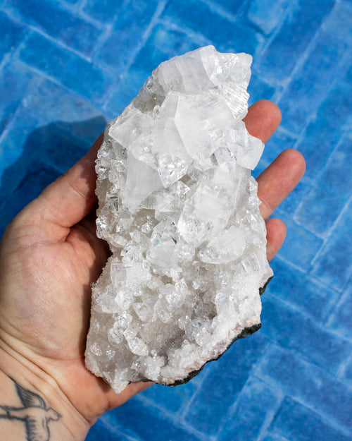 A hand holding a large, rough white crystal cluster with clear faceted mineral pieces, set against a blue tiled background.