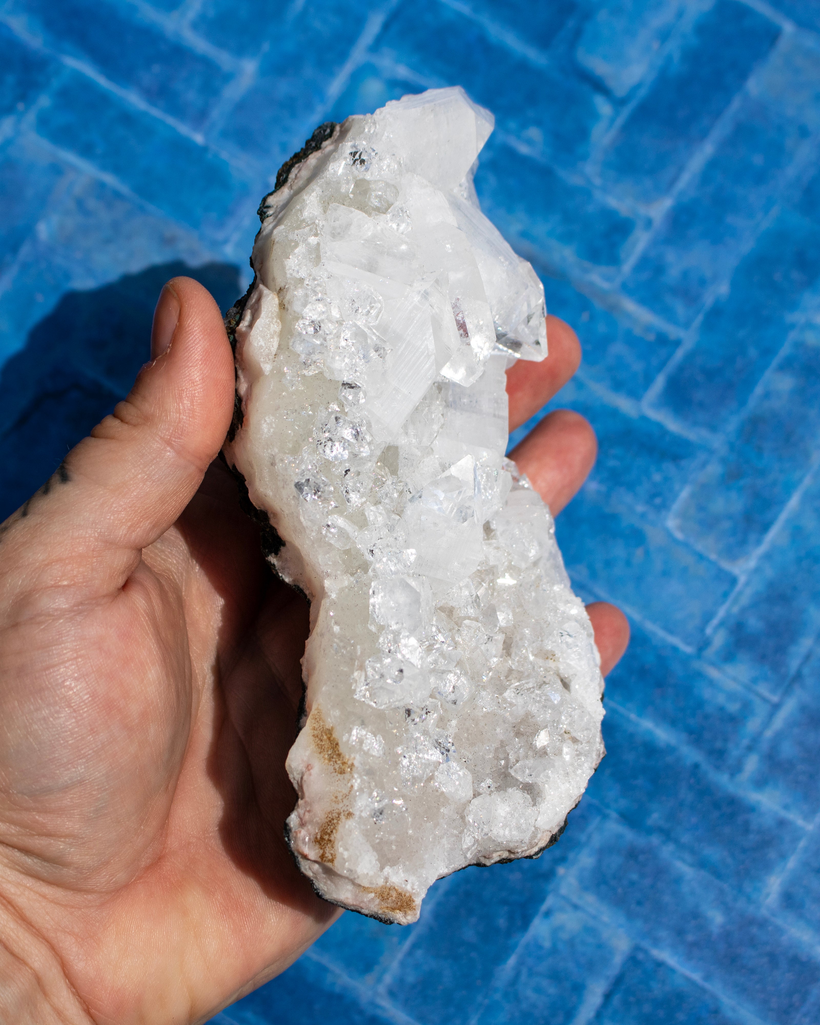 A hand holding a raw crystal cluster with clear quartz-like formations and a rough white to gray matrix, photographed against a blue tiled background.