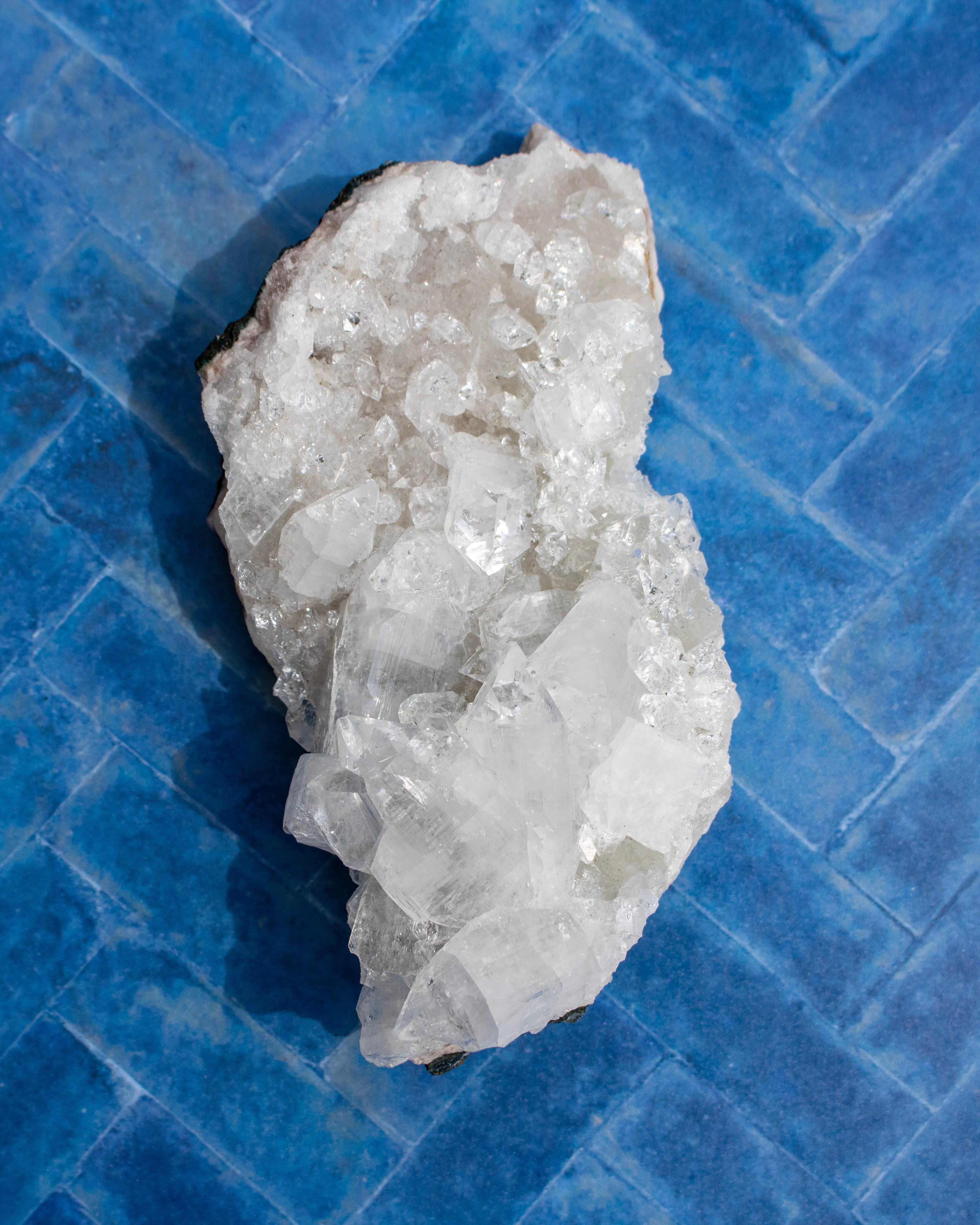 A raw white quartz crystal cluster with rough, natural edges and a dark matrix, displayed on a blue tiled surface.