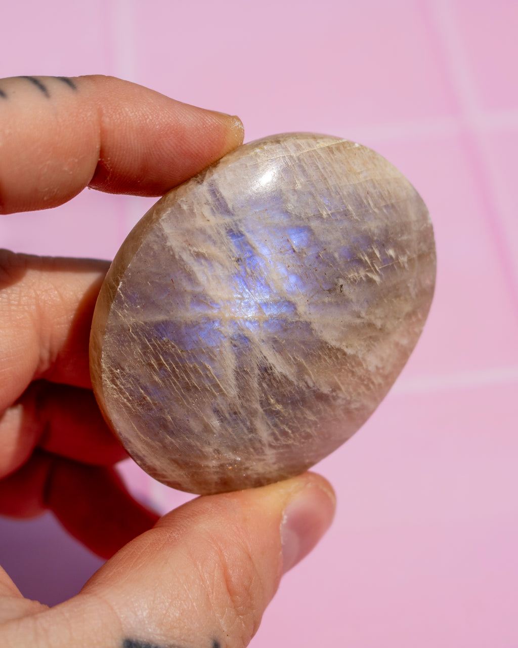 A hand holding a polished oval stone with a lavender-purple sheen and fine crystalline texture against a pink background.