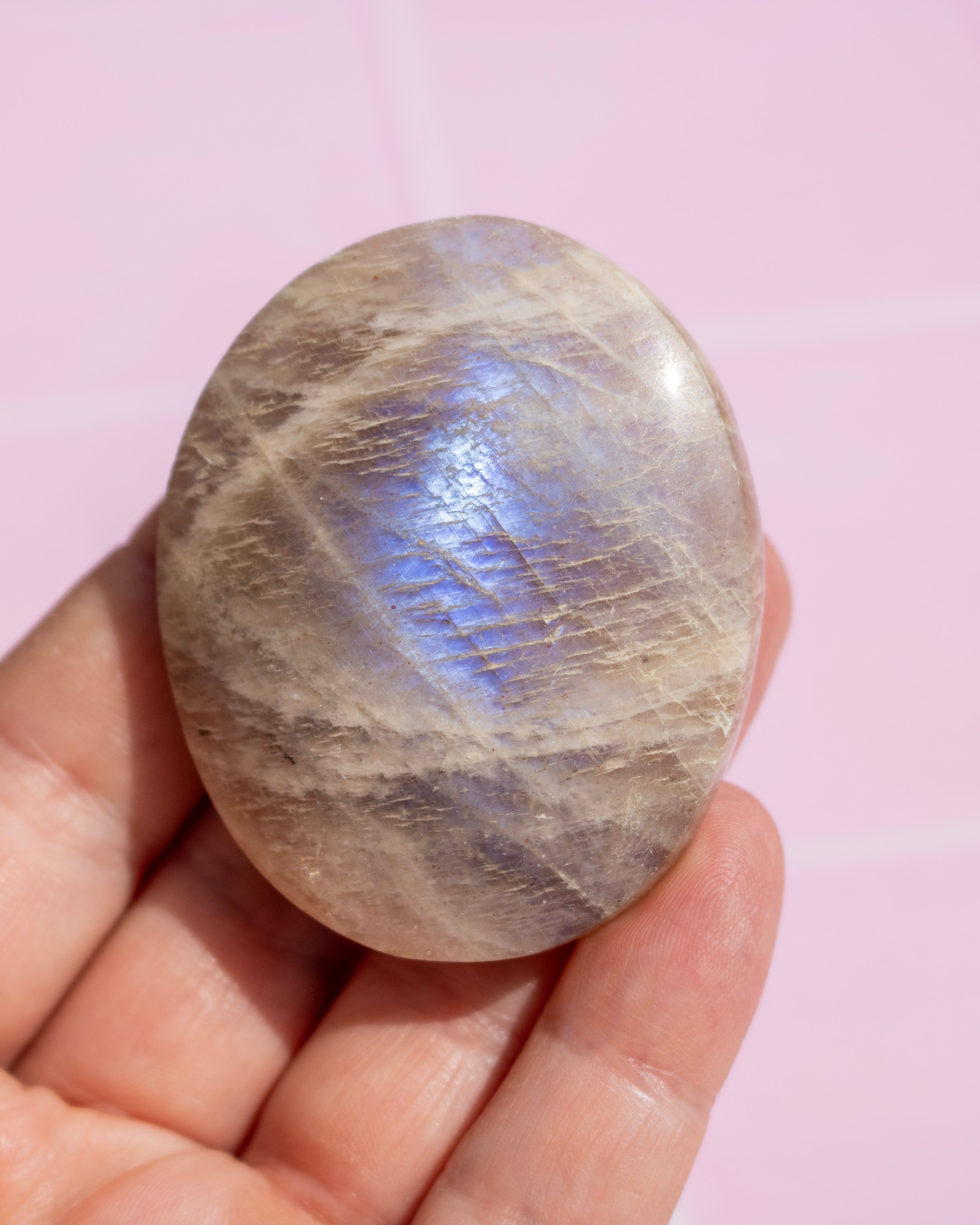 A hand holding a polished oval stone with a soft iridescent sheen, showing beige-gray tones with blue and purple highlights, against a pale pink background.