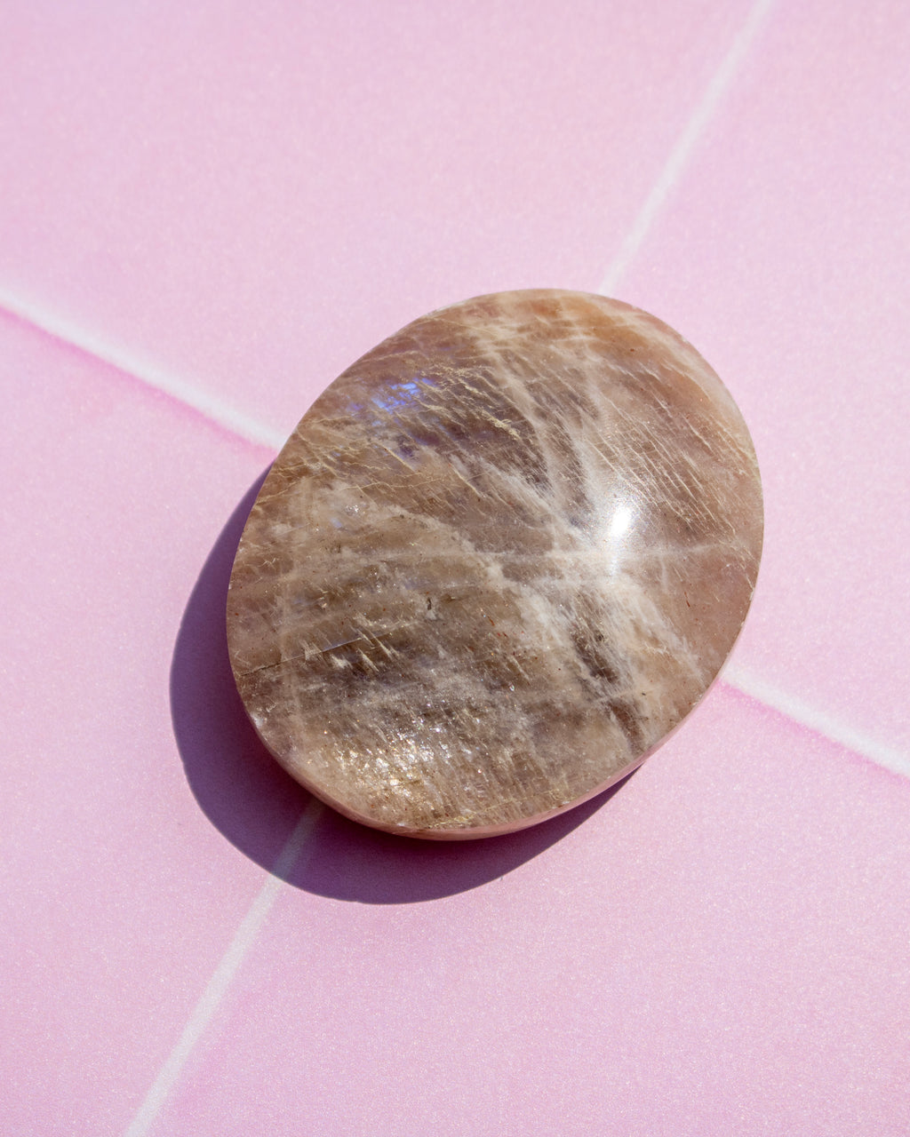 A polished oval-shaped brown gemstone with fine white and gray crystalline striations, resting on a pink background with a soft shadow.