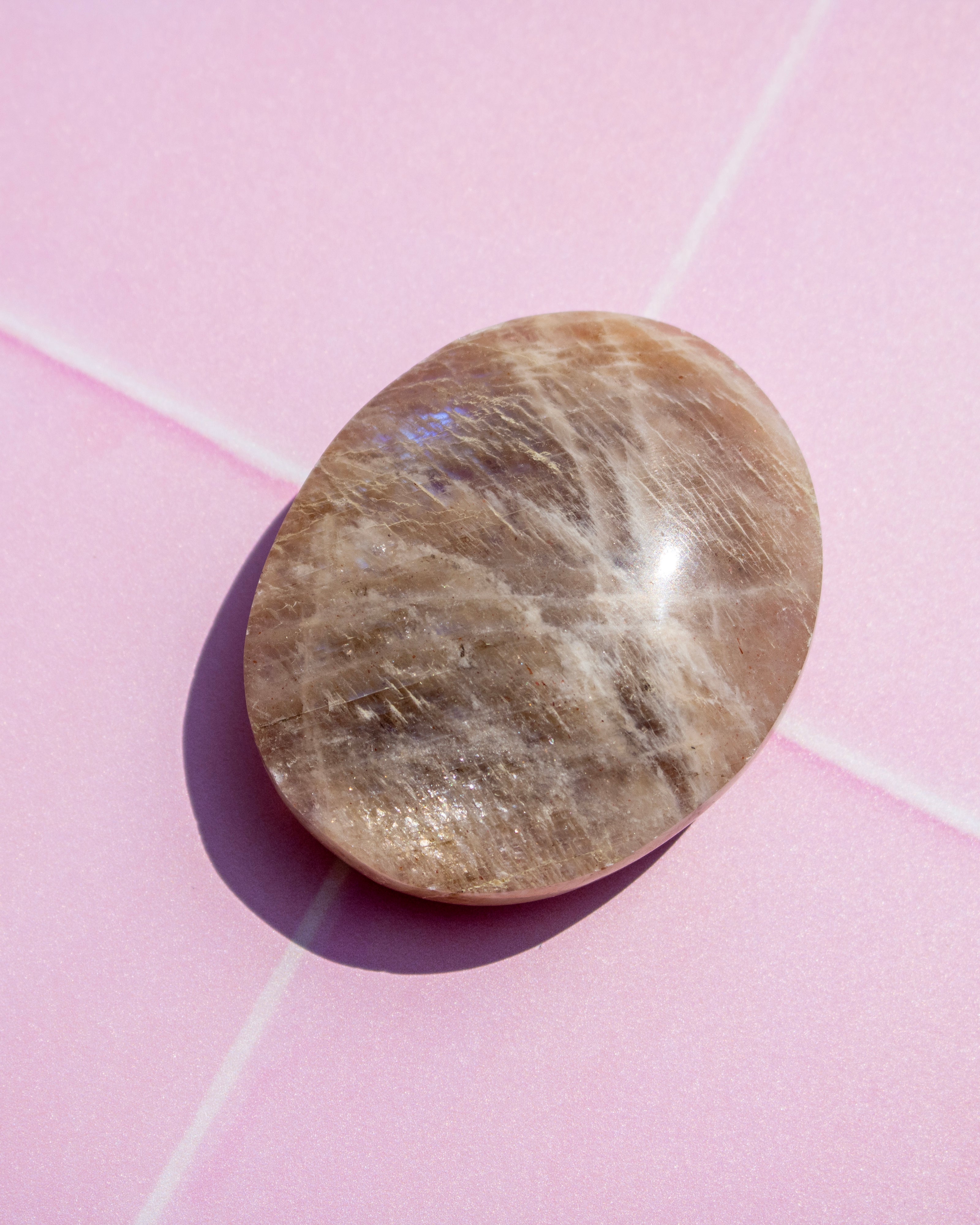 A polished oval-shaped brown gemstone with fine white and gray crystalline striations, resting on a pink background with a soft shadow.