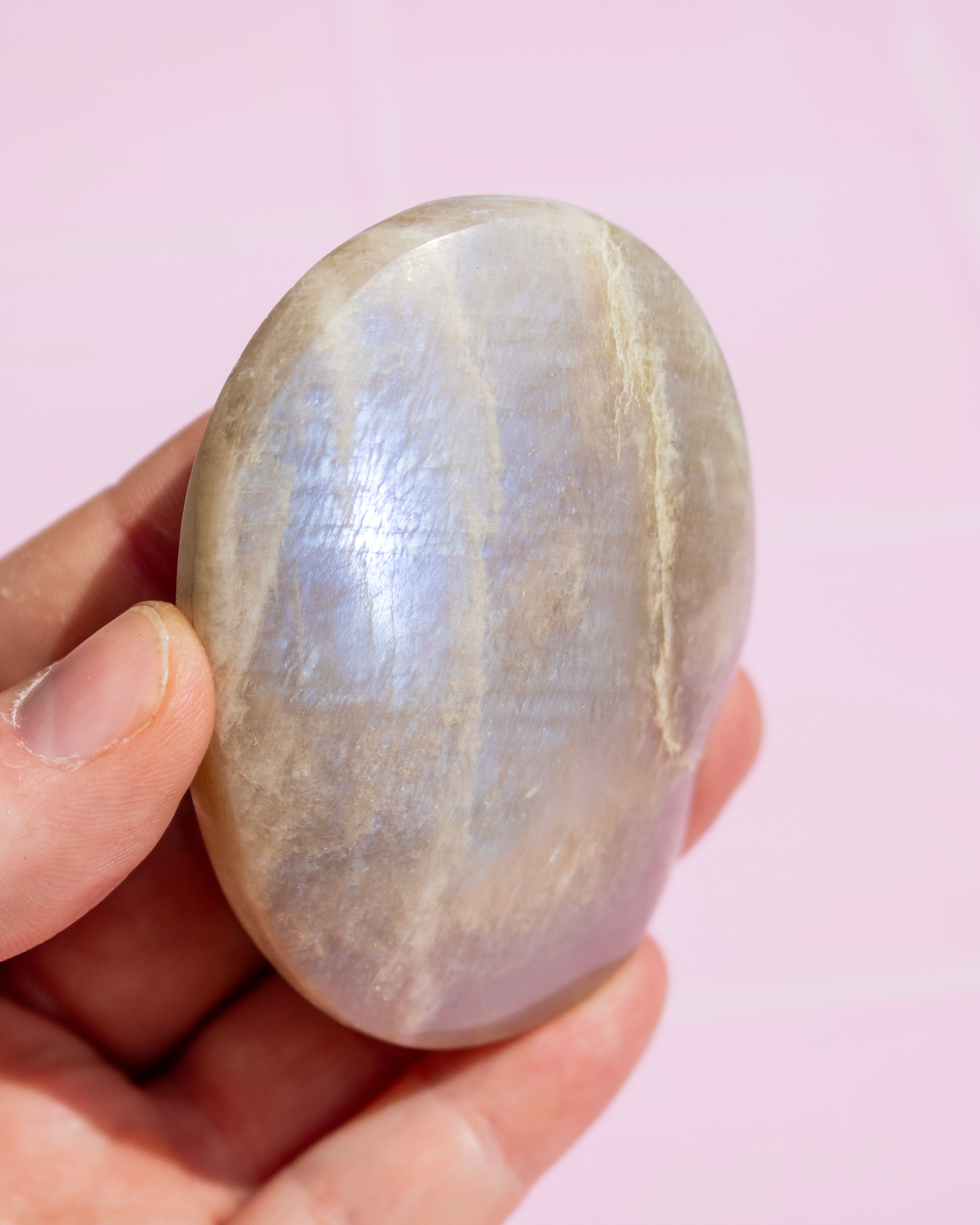 A hand holding a polished oval stone with a pearlescent lavender-gray sheen against a soft pink background.