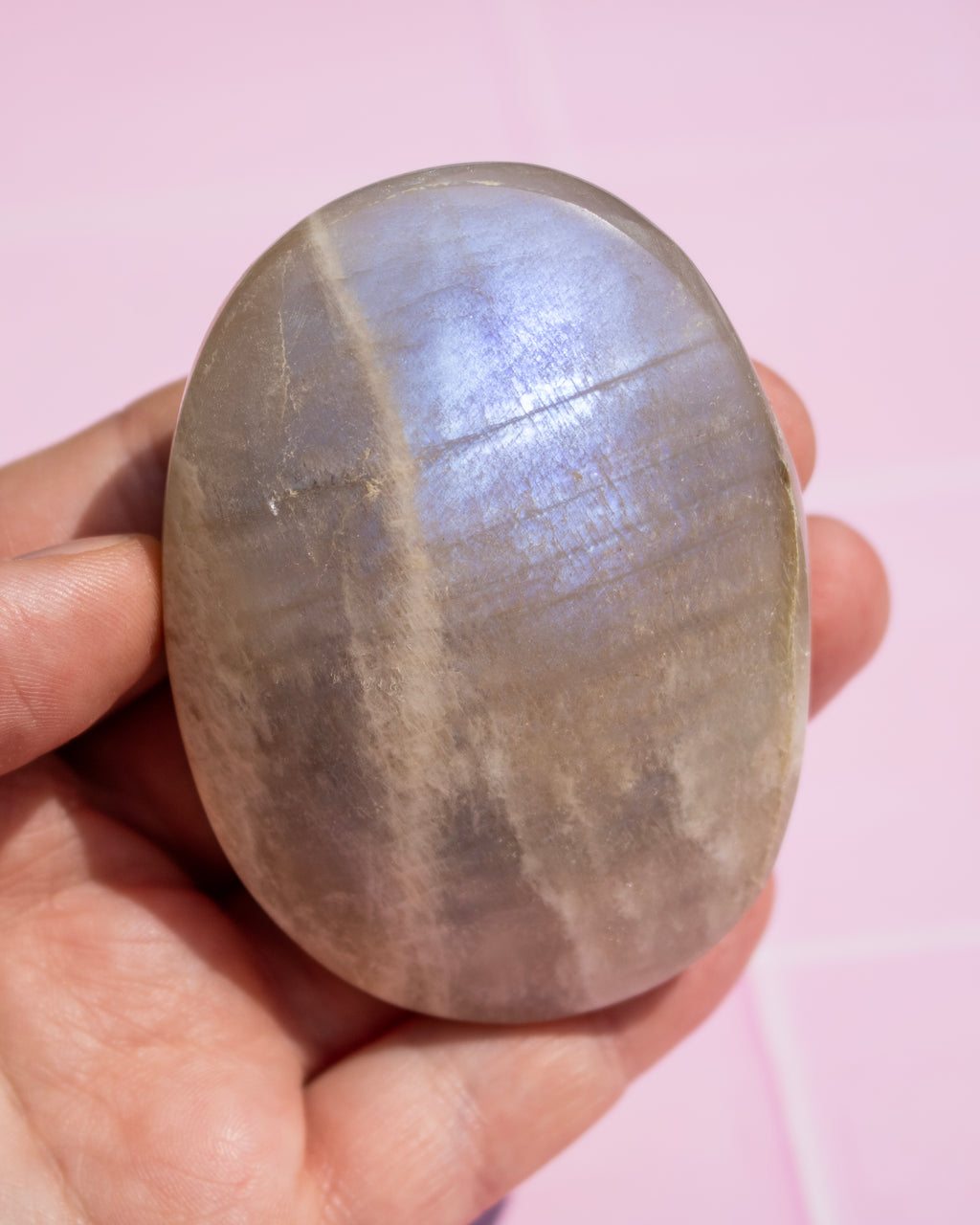 A hand holding a smooth, polished oval stone with a metallic silver-gray luster, set against a soft pink background.