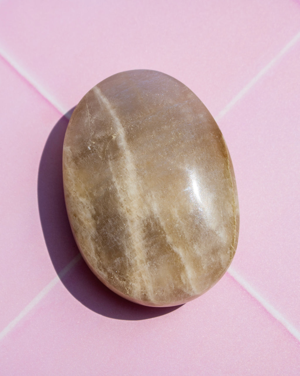 A polished oval stone with natural beige and light brown veining, resting on a pink background with a soft shadow.