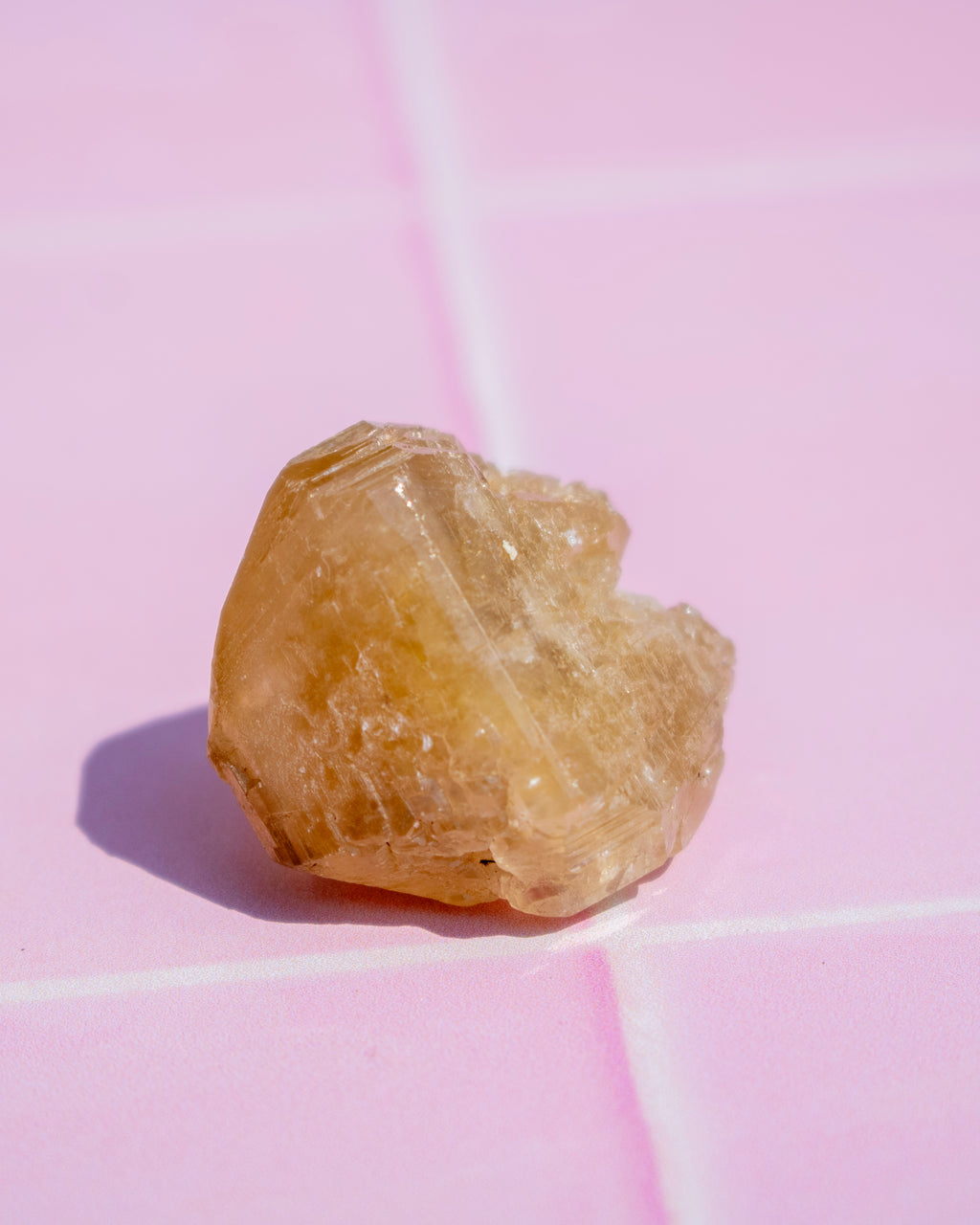 A small rough, translucent yellowish-brown crystal or gemstone photographed on a pink surface with visible grid lines and a cast shadow.