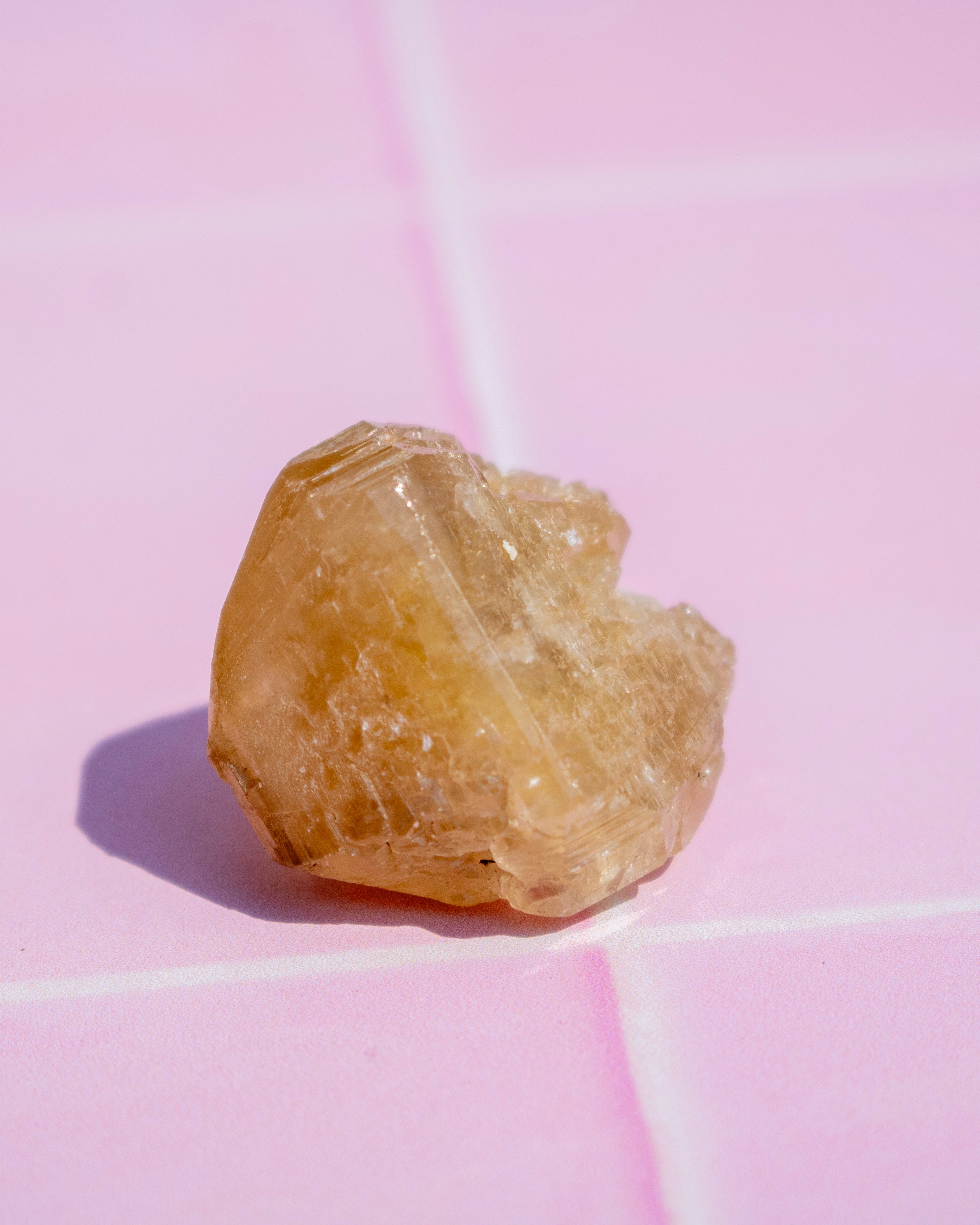A small rough, translucent yellowish-brown crystal or gemstone photographed on a pink surface with visible grid lines and a cast shadow.