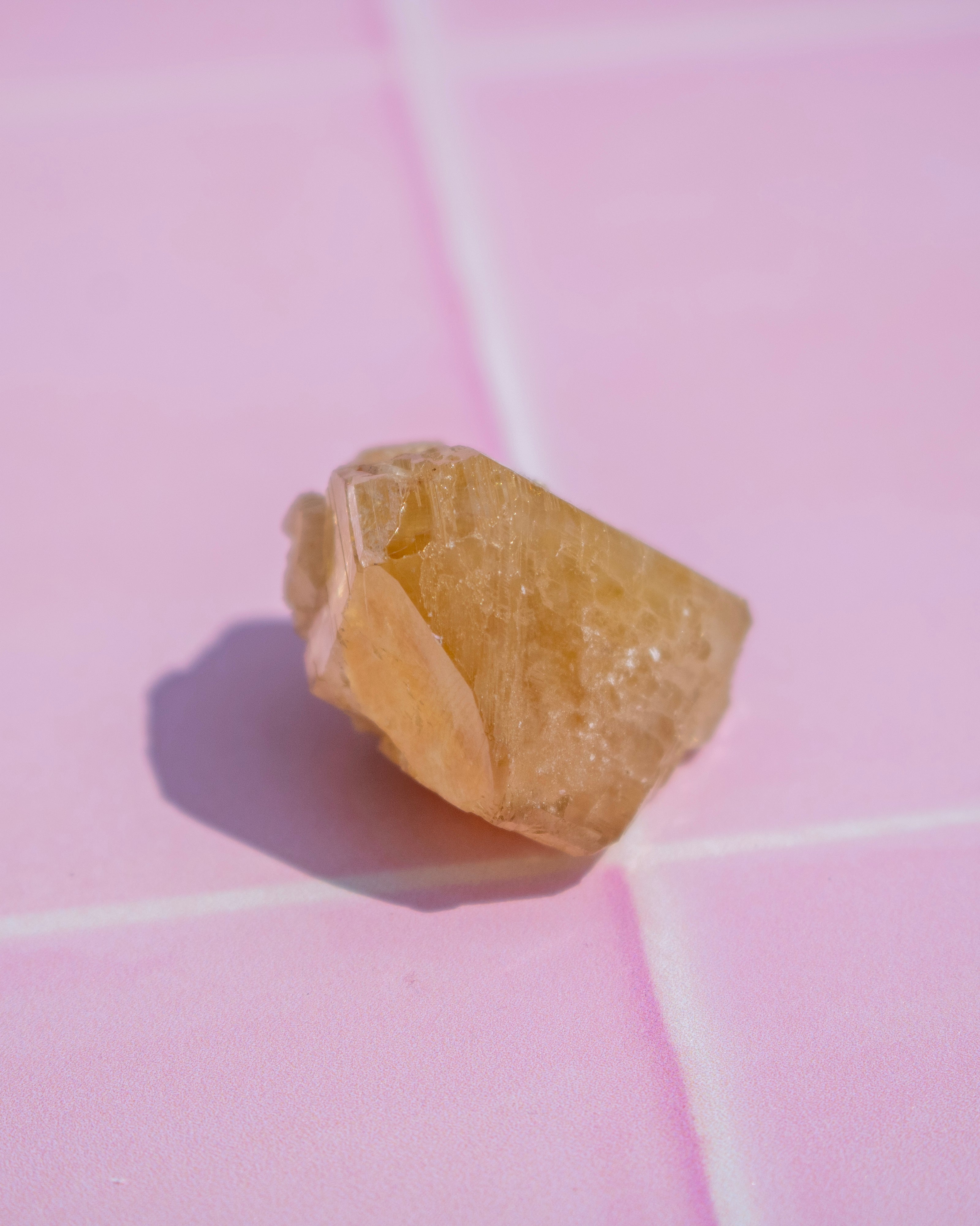 A rough, unpolished yellow crystal or gemstone resting on a pink tiled surface, casting a soft shadow to the left.