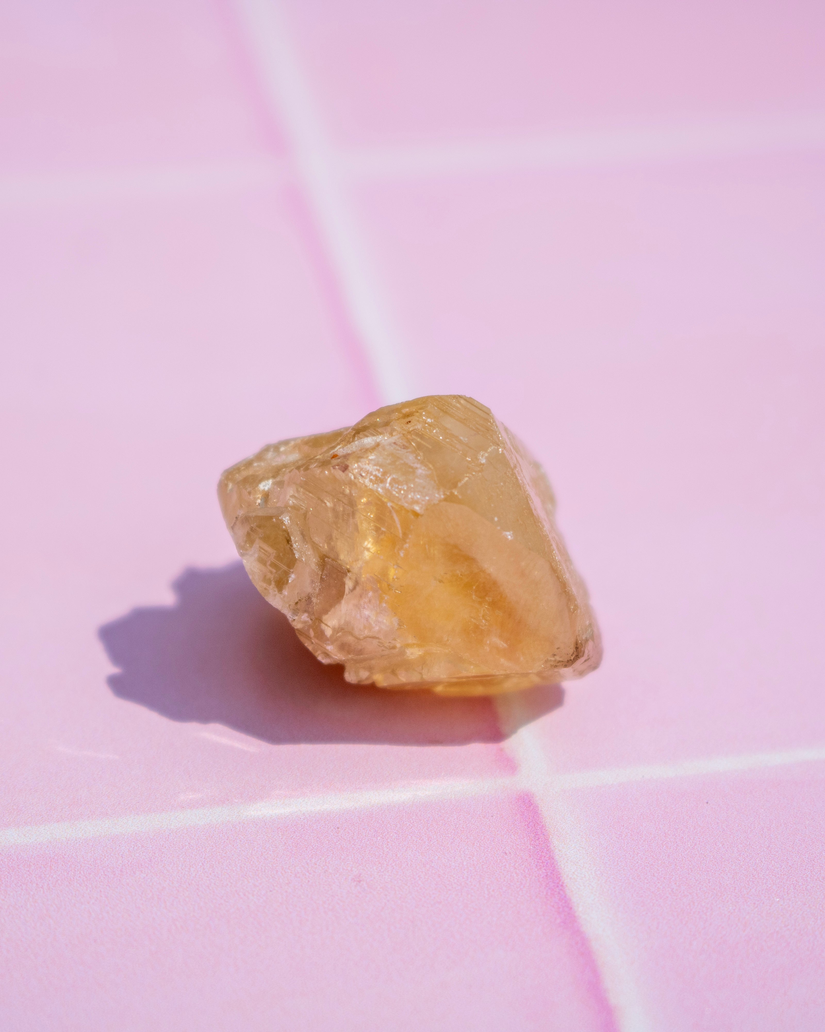 A small, rough, translucent yellow crystal or gemstone resting on a pink tiled surface with white grout lines.