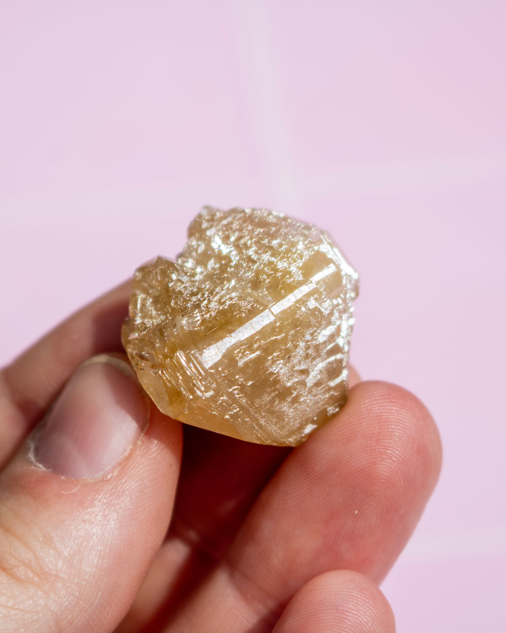 A hand holding a rough, unpolished yellow crystal or gemstone against a soft pink background.