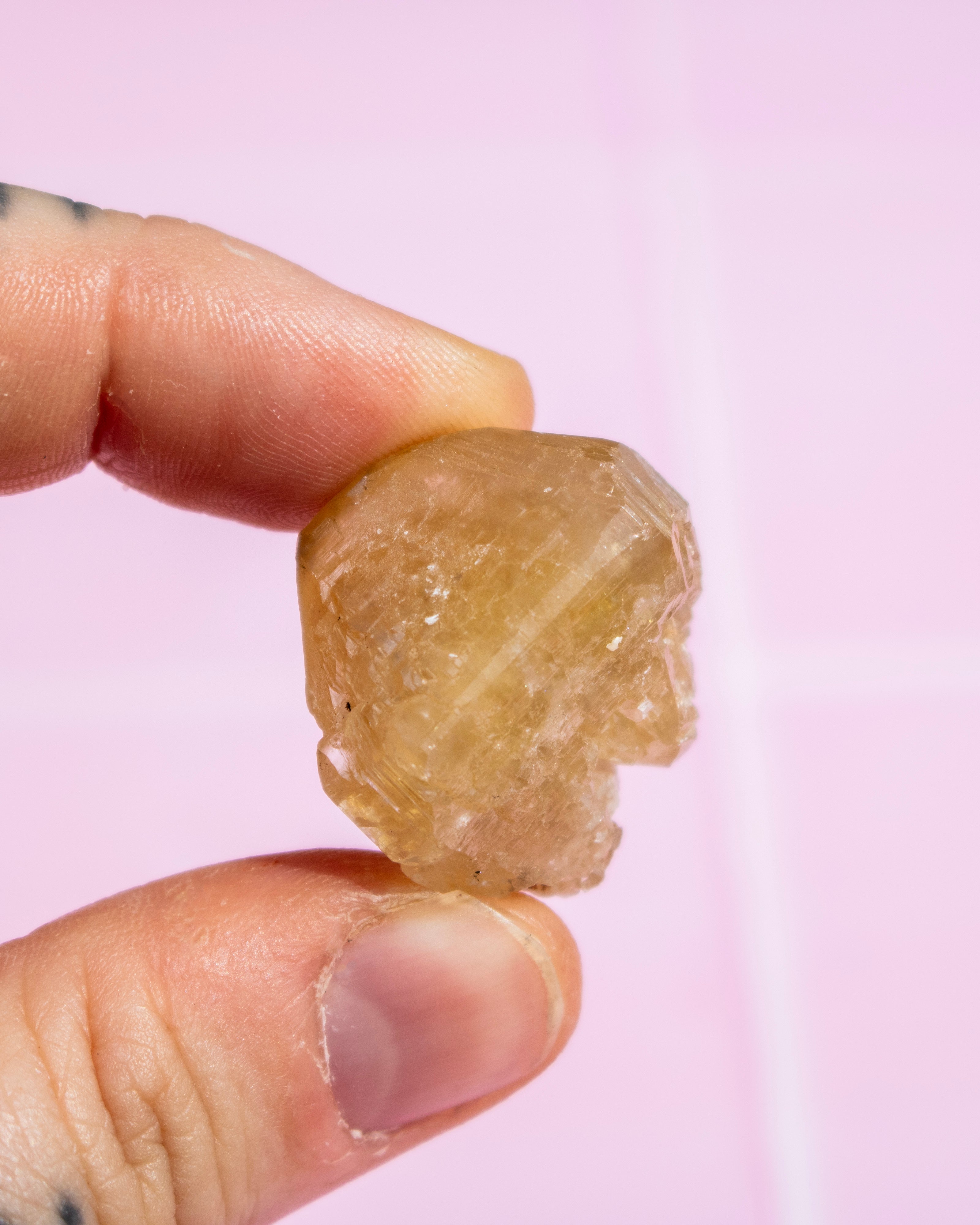 A hand holding a small, rough, translucent pale yellow crystal or gemstone against a soft pink background.