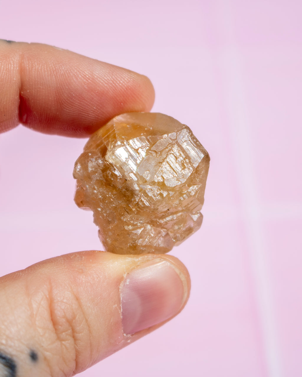 A hand holding a small, rough, translucent golden-brown crystal or mineral specimen against a soft pink background.