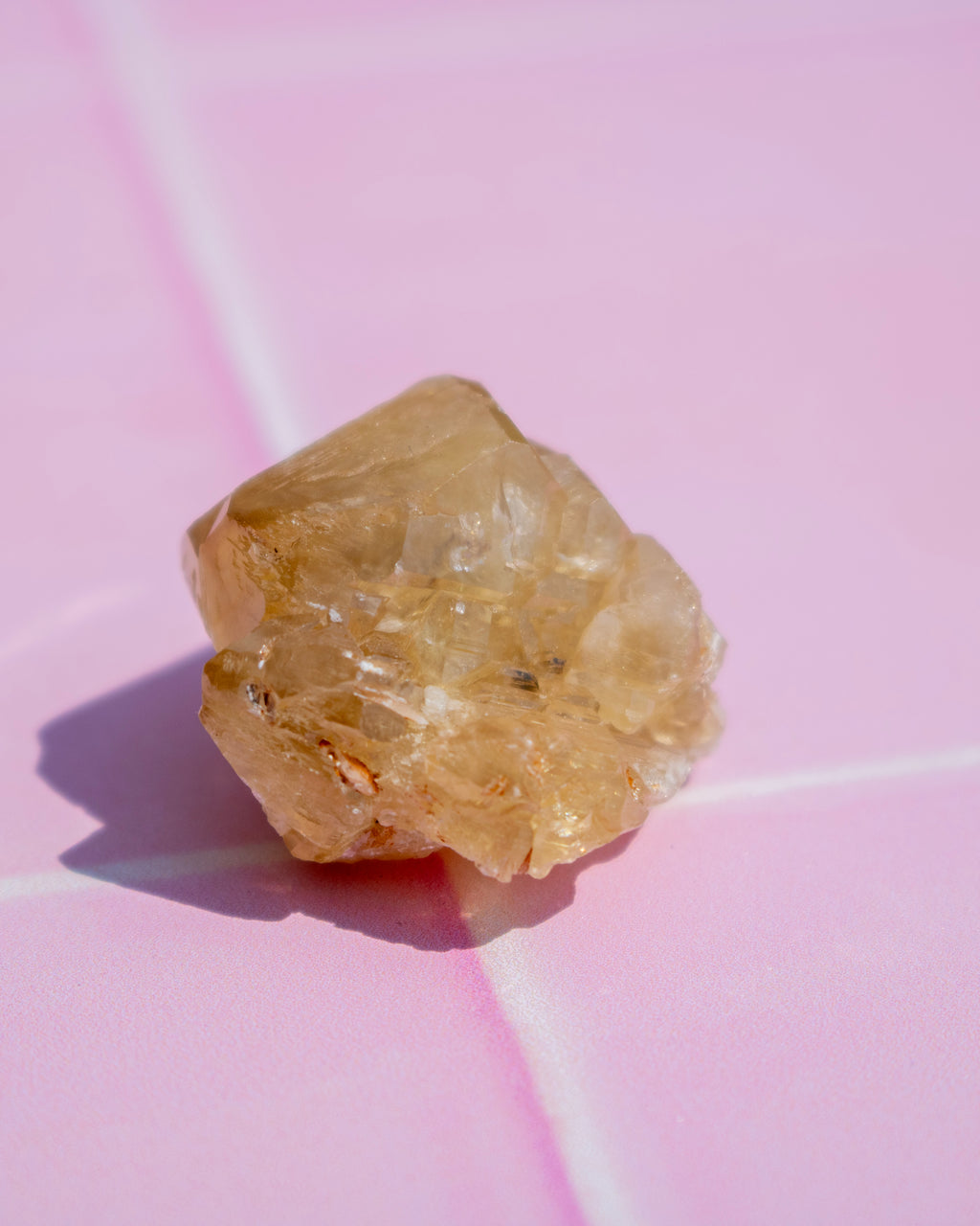 A rough, translucent pale yellow crystal or mineral specimen resting on a pink surface with white grout lines.