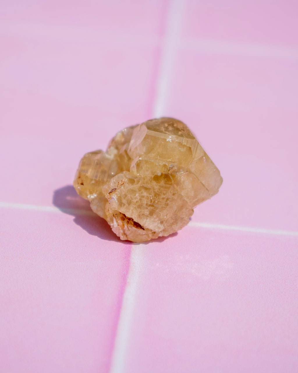 A small, rough, pale yellow translucent crystal cluster resting on a pink tiled surface.