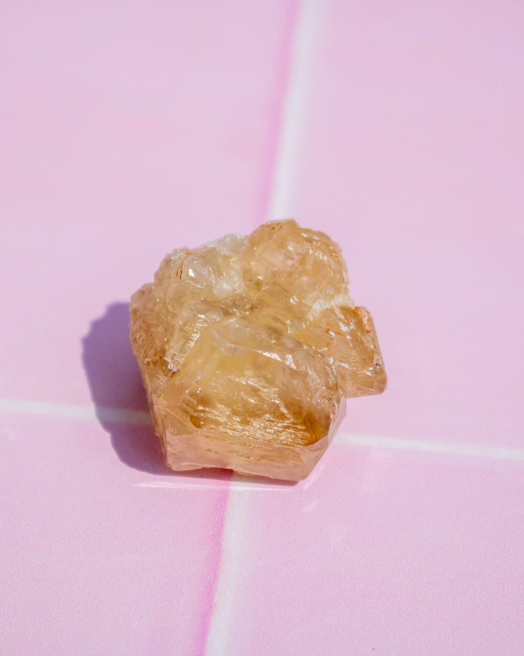 A rough, unpolished piece of translucent yellow stone or crystal, resembling citrine, resting on a glossy pink surface with a soft shadow cast to the left.