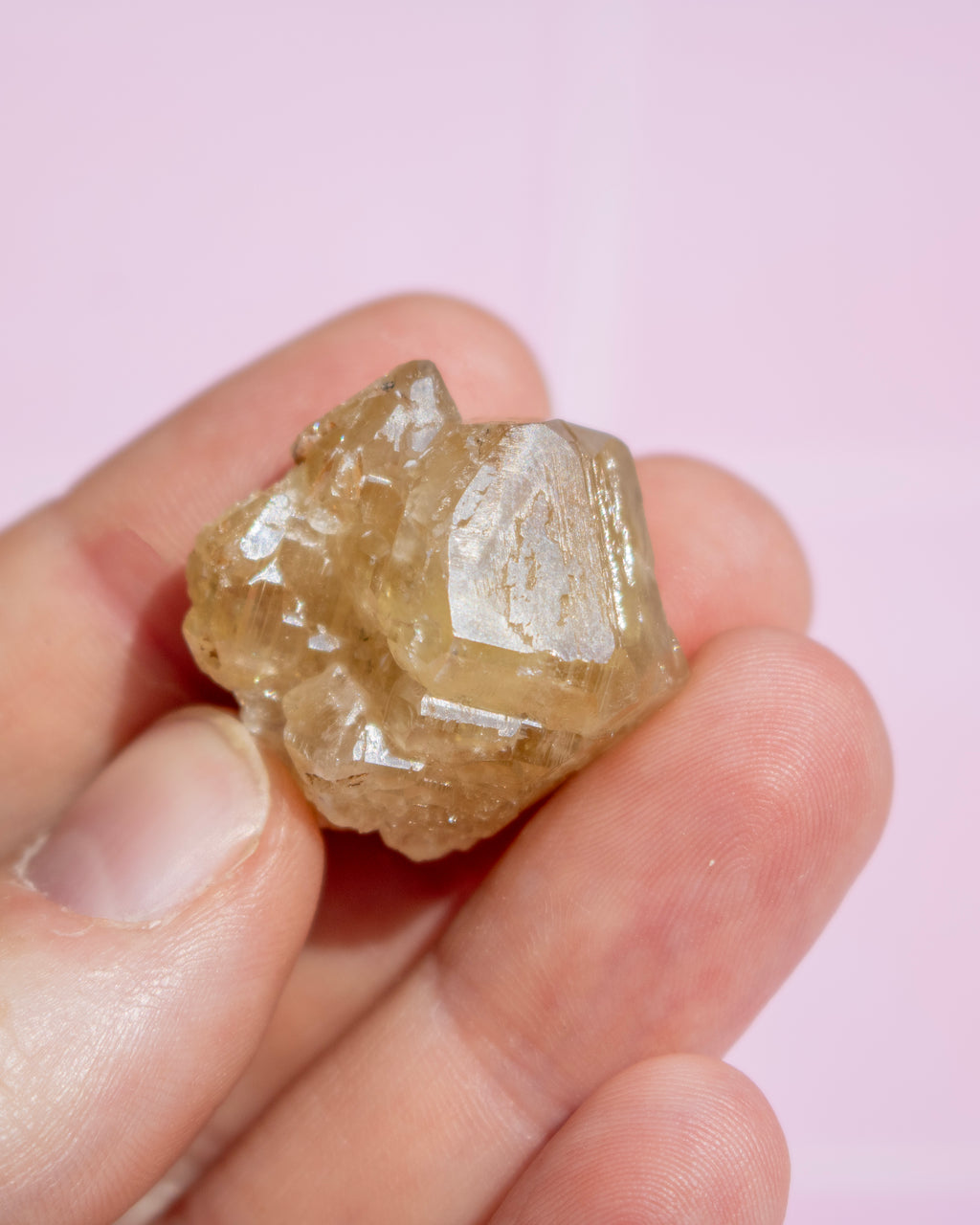 A hand holding a small, rough, translucent yellowish-brown crystal against a soft pink background.