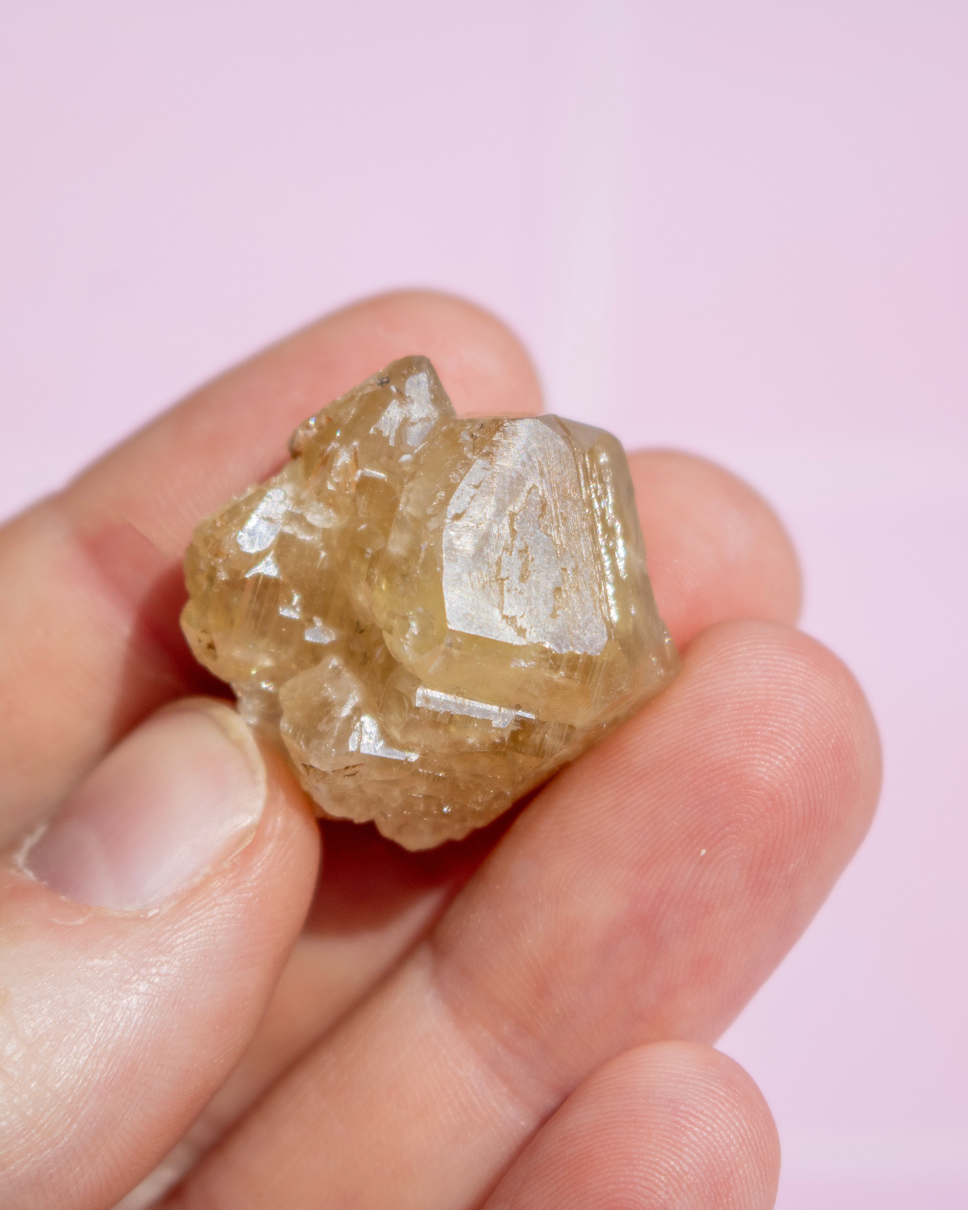 A hand holding a small, rough, translucent yellowish-brown crystal against a soft pink background.