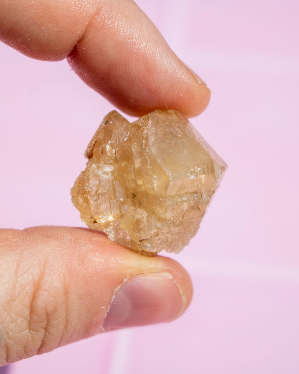 A close-up of a hand holding a small, rough, translucent pale yellow crystal against a soft pink background.