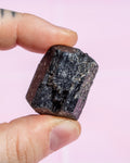 A close-up of a hand holding a rough, dark polished mineral specimen against a soft pink background.