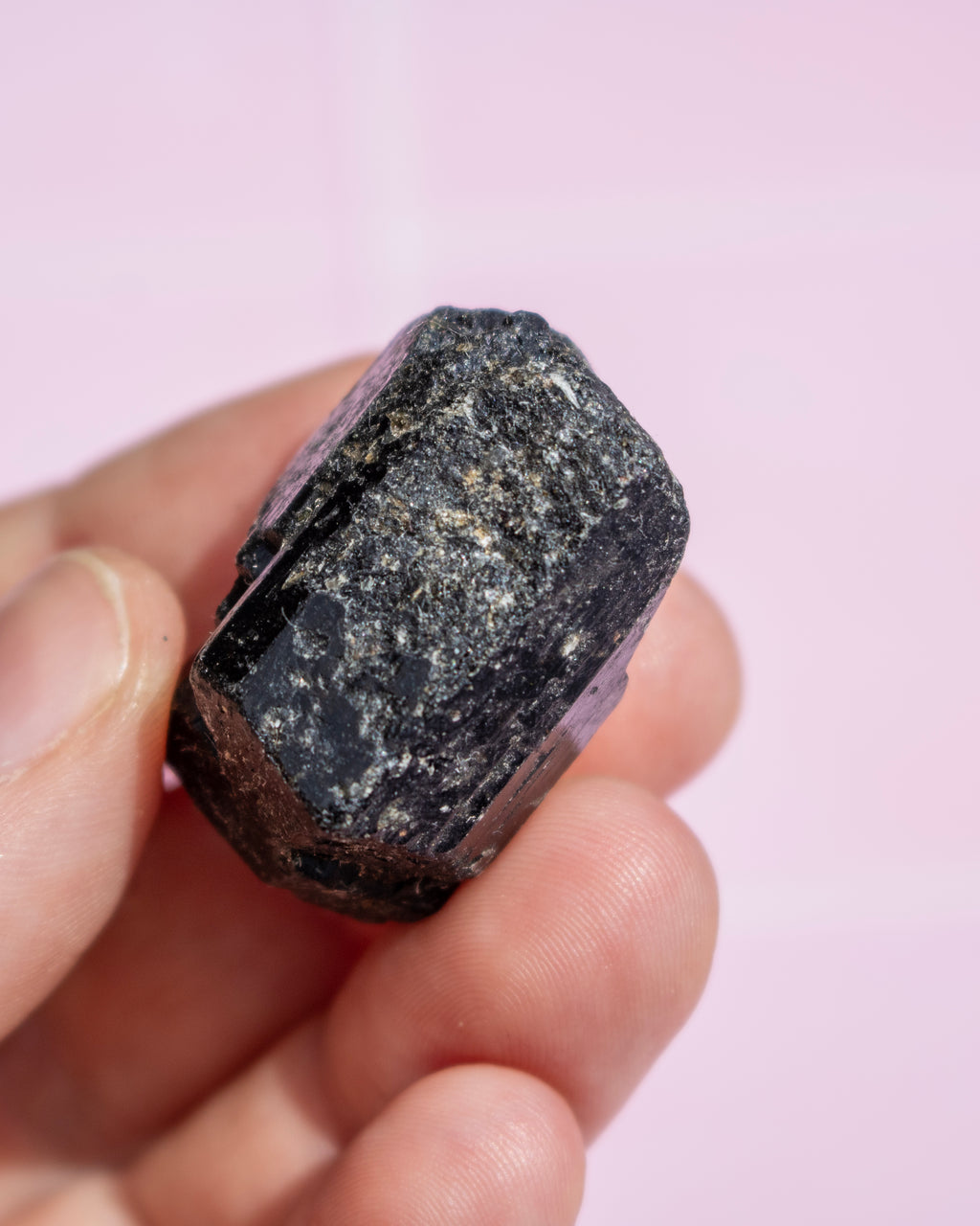 A hand holding a rough, dark polished crystal or mineral specimen against a soft pink background.