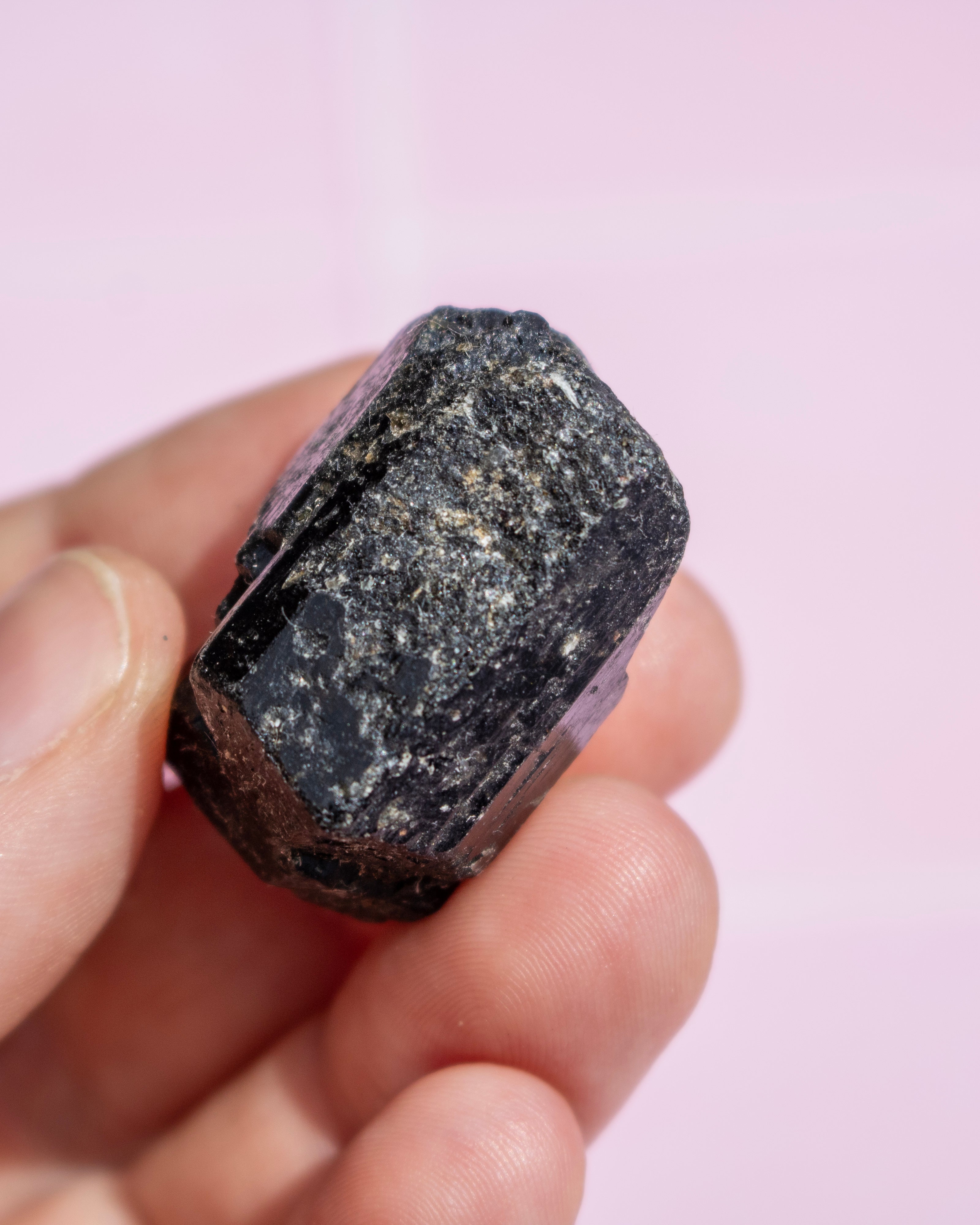 A hand holding a rough, dark polished crystal or mineral specimen against a soft pink background.