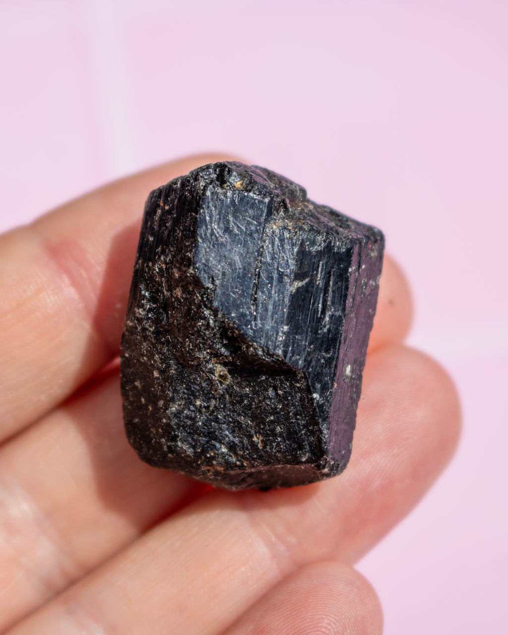 A rough, dark polished mineral specimen held in a person's hand against a soft pink background.