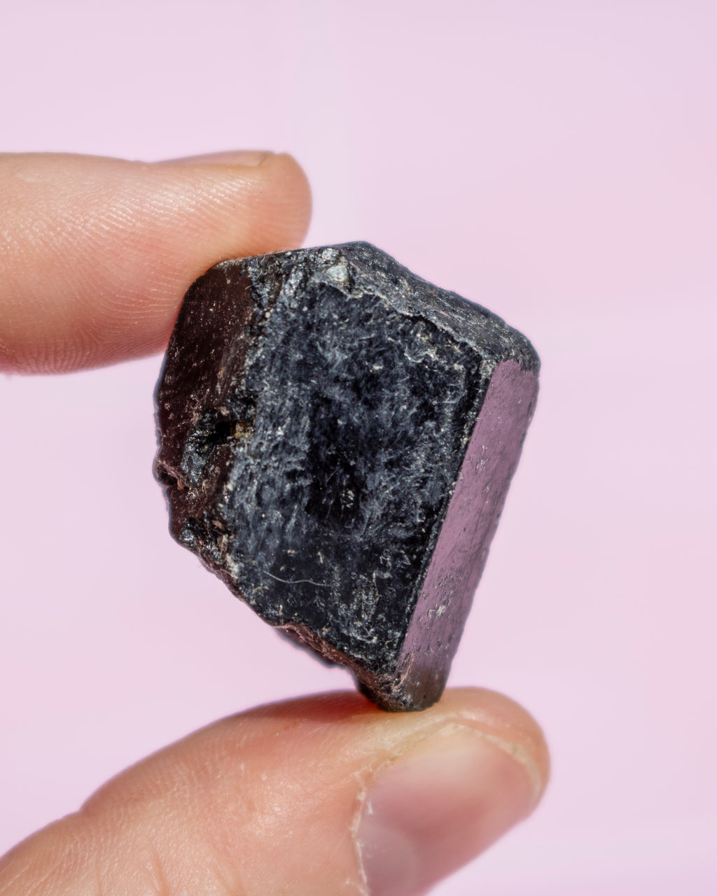 A hand holding a rough, dark polished crystal with a hexagonal shape against a solid light pink background.
