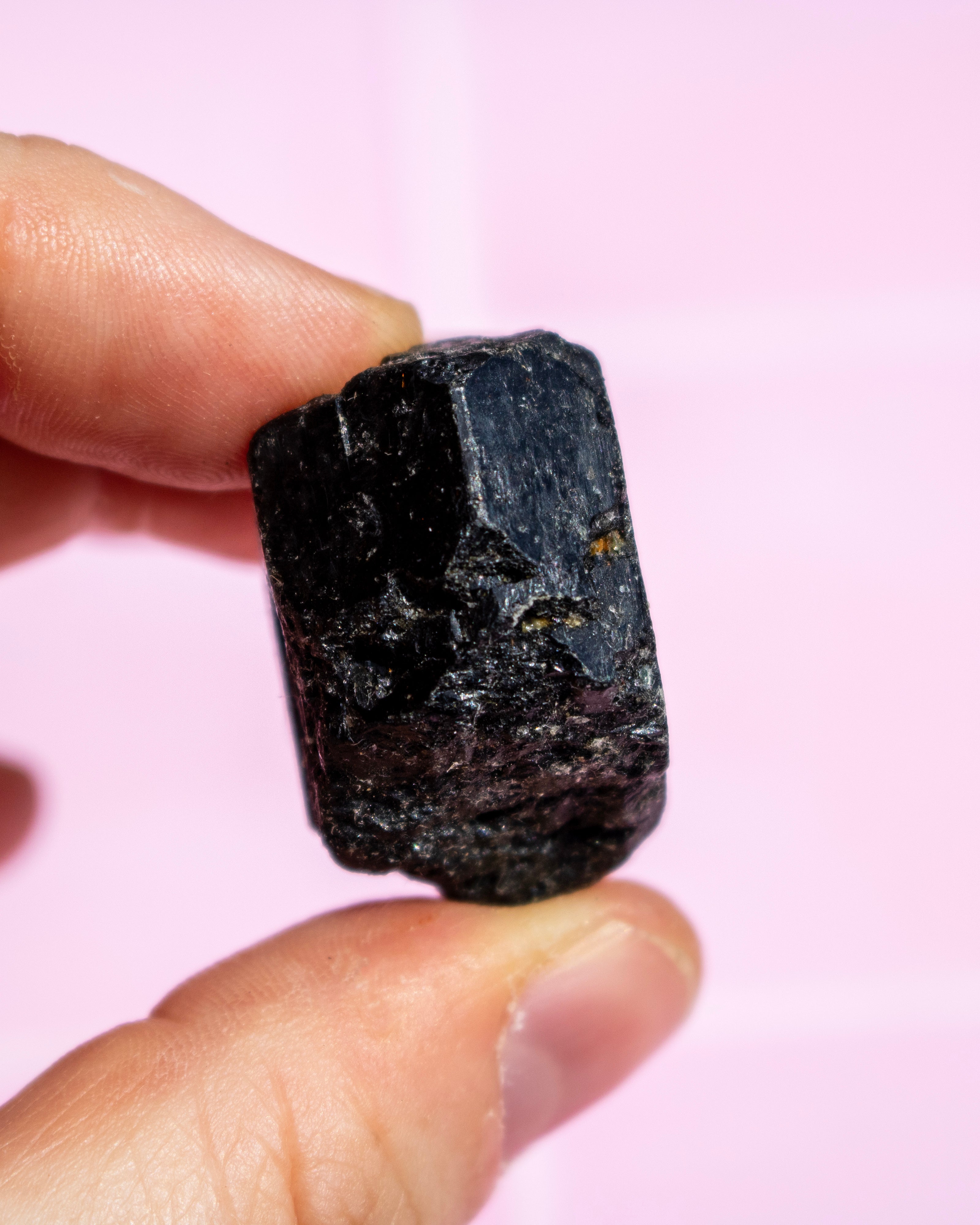A hand holding a small, rough, dark black mineral or crystal against a soft pink background.