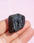 A rough, dark natural mineral or gemstone held on a person's fingers against a soft pink background.