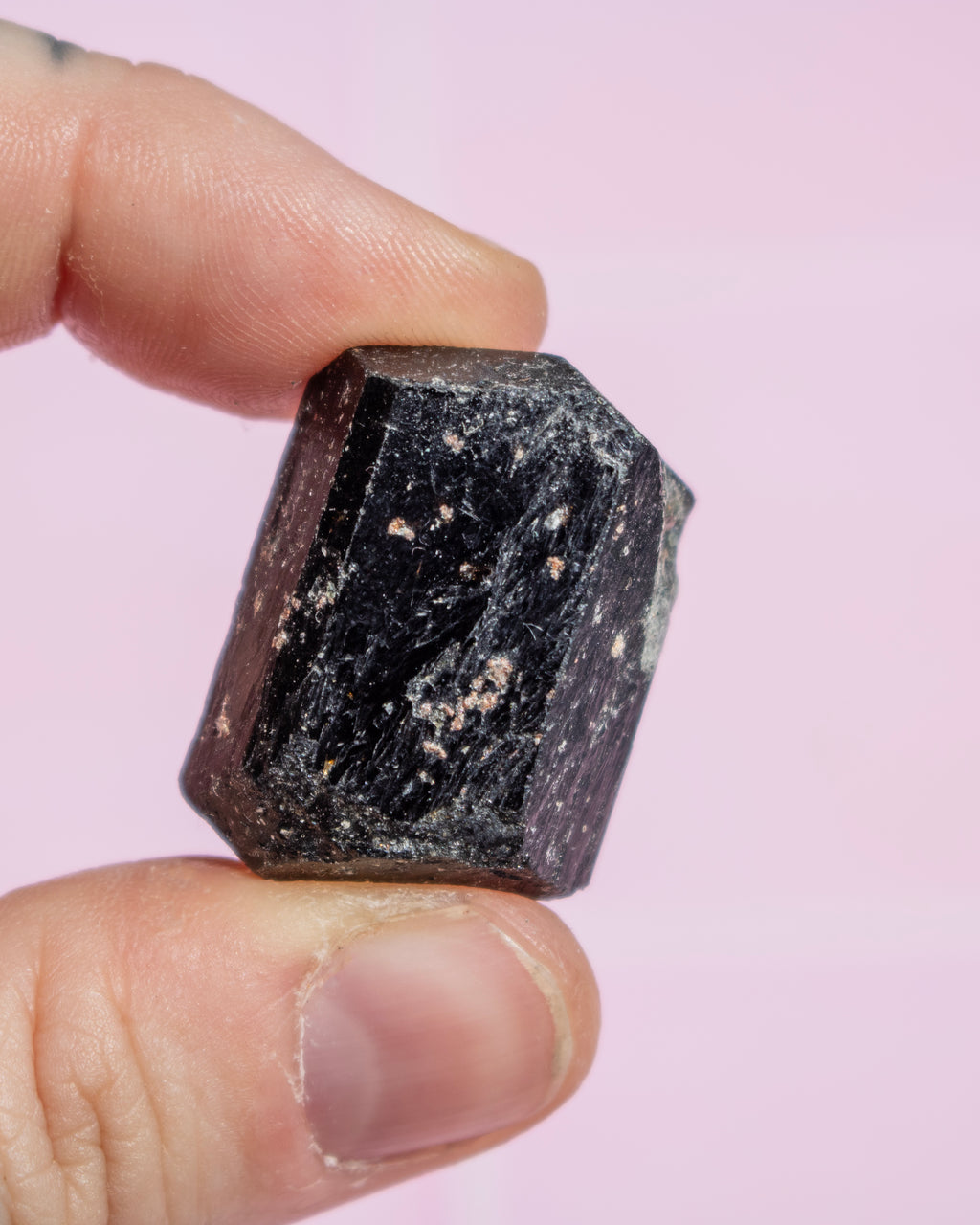 A hand holding a small faceted dark mineral or crystal against a soft pink background.