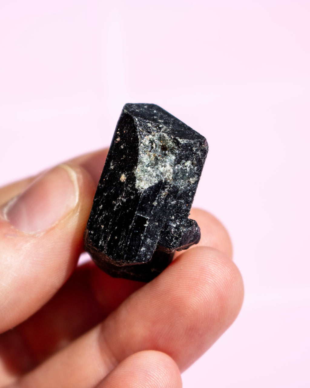 A hand holding a small, rough black mineral or crystal specimen against a soft pink background.
