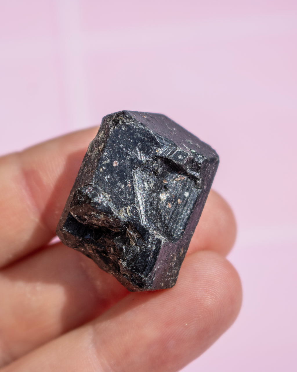 A hand holding a rough, faceted black gemstone or mineral against a soft pink background.