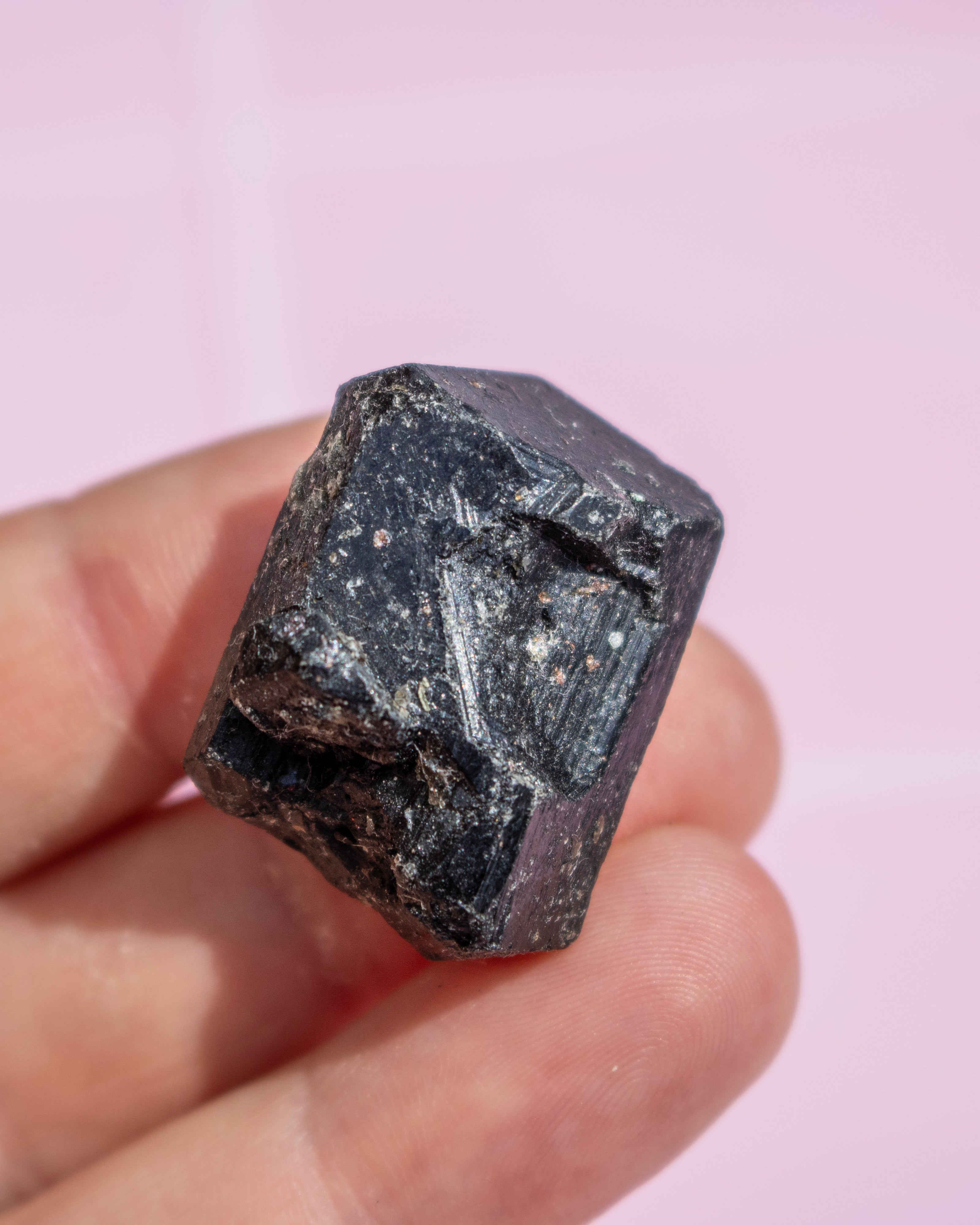 A hand holding a rough, faceted black gemstone or mineral against a soft pink background.