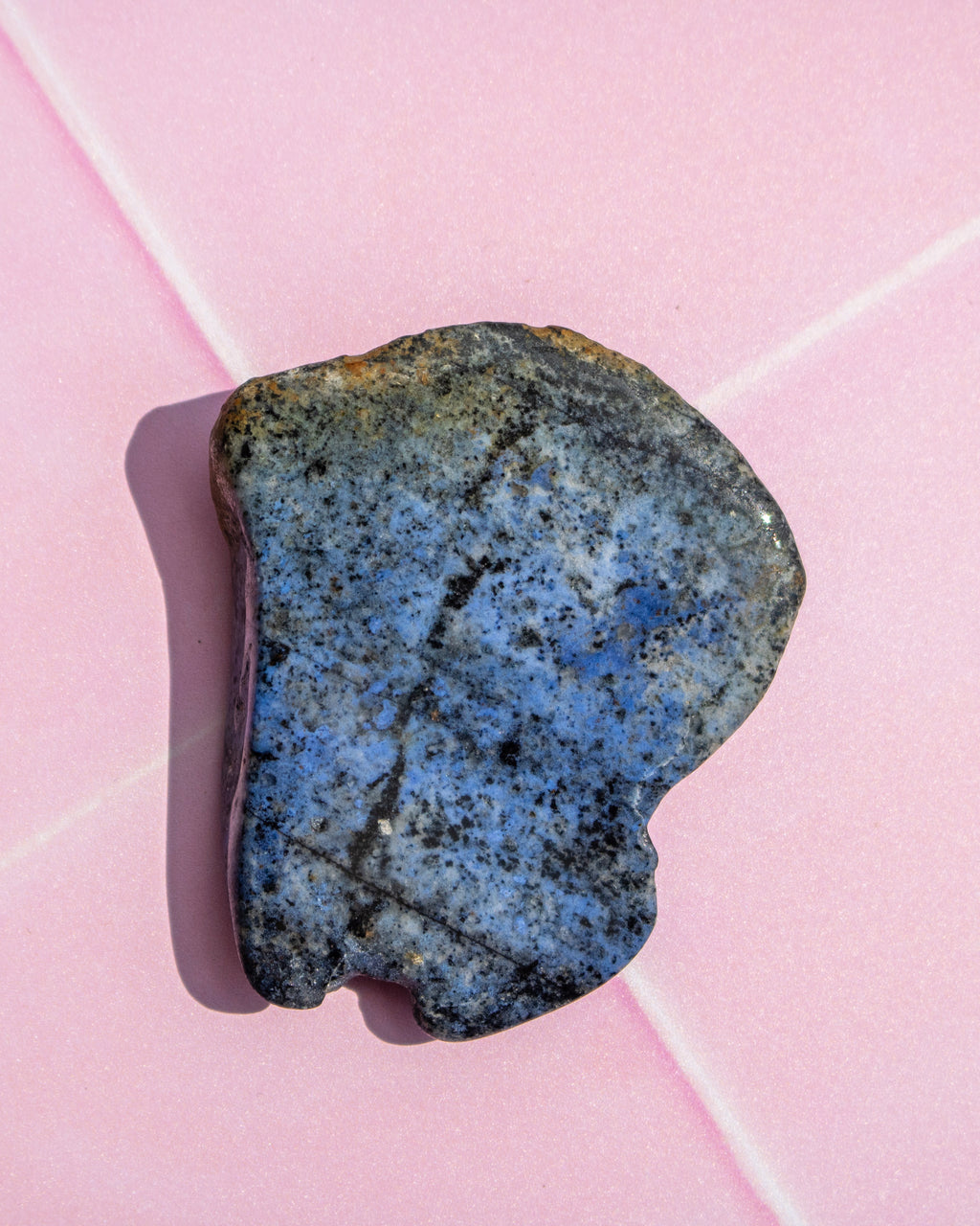 A polished blue stone with black and gray speckles, resting on a light pink background with white diagonal lines.