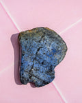 A polished blue stone with black and gray speckles, resting on a light pink background with white diagonal lines.