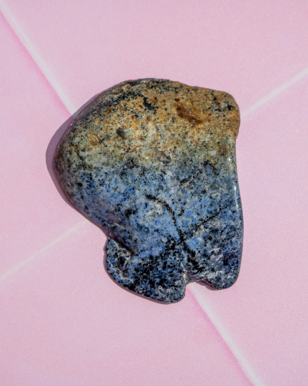A polished natural stone with a mottled color transition from tan and gray at the top to blue and black at the bottom, resting on a light pink background.